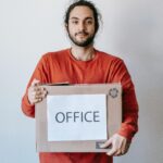 Smiling young man holding a cardboard box labeled 'Office', implying relocation.